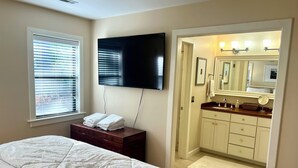 A bedroom with a bed, a TV mounted on the wall, a window with blinds, and a bathroom visible through an open door.
