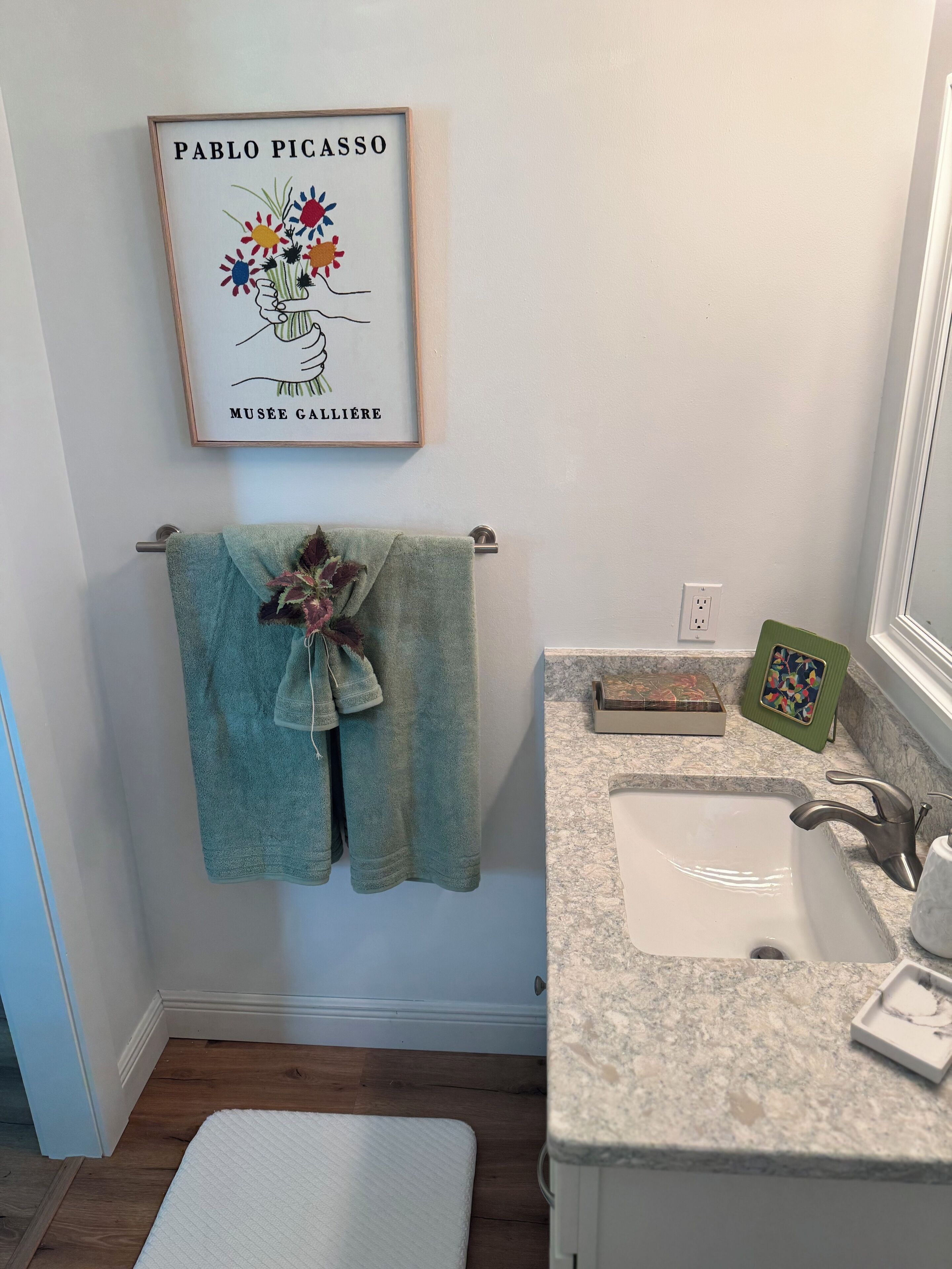 Een badkamer met een afbeelding van Pablo Picasso in een lijst, een groene handdoek, een witte badmat, een wastafel en een spiegel.