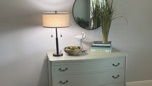 Eine hellblaue Kommode mit drei Schubladen, ein runder Spiegel, eine Lampe, eine Schüssel mit grünen Äpfeln, eine weiße Vogel-Skulptur und ein Stapel Bücher.