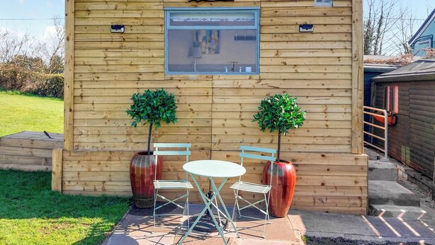 A wooden cabin labeled "Sunset Cabin" with a small table and chairs, two potted plants, and a window. - 1 bedroom accommodation in Ormskirk (Ormskirk)