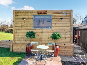Una cabaña de madera con un letrero que dice 'Sunset Cabin', una pequeña mesa y sillas, dos plantas en macetas y un ventanal.