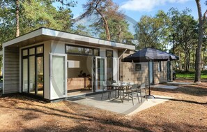 Una cabaña moderna con puerta de cristal, un patio con mesa y sillas, y una zona de estar cubierta.