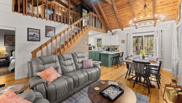 A spacious living area with a leather sofa, a wooden staircase, a dining table with chairs, and a kitchen in the background.