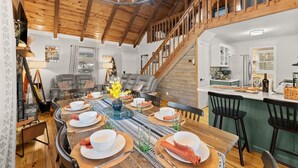A dining area with a wooden table set for six, a staircase, and a kitchen in the background.