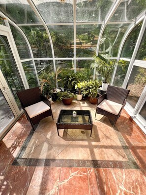A conservatory with a glass ceiling, two wicker chairs with cushions, a glass coffee table, and various potted plants. - US Open Stay - Hudson River View Estate with Private Tennis Court (Nyack)