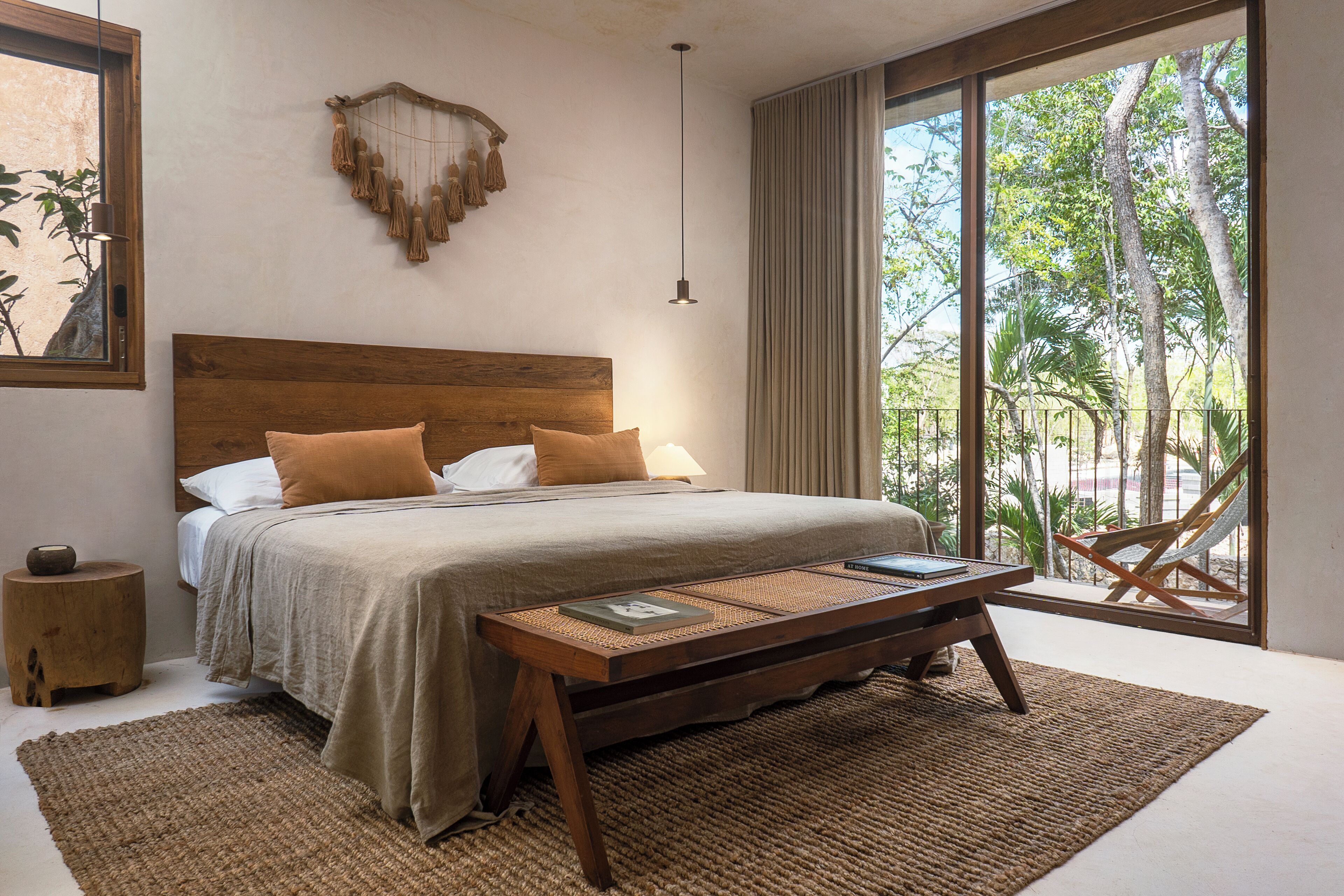 A bedroom with a wooden headboard, a bed with beige bedding, a woven side table, and a large window with a view of trees.