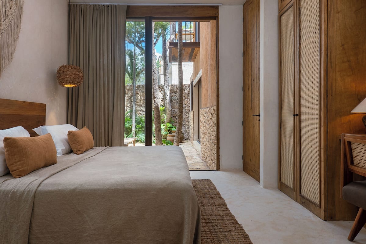 A bedroom with a large bed, a wooden headboard, a woven light fixture, and a view of a stone wall and greenery outside.