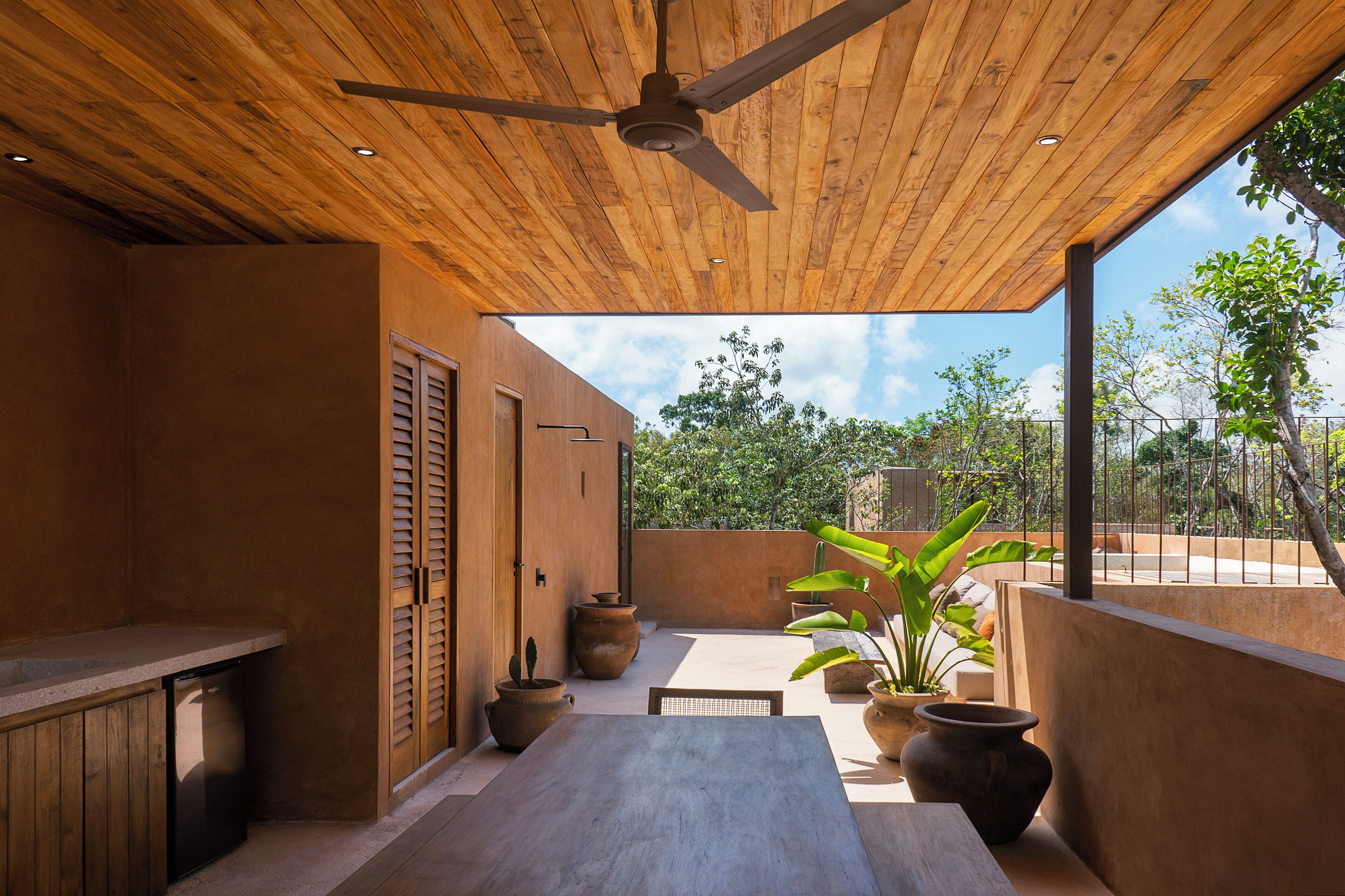 An outdoor patio with a wooden ceiling, a dining table, potted plants, and a view of the surrounding greenery.