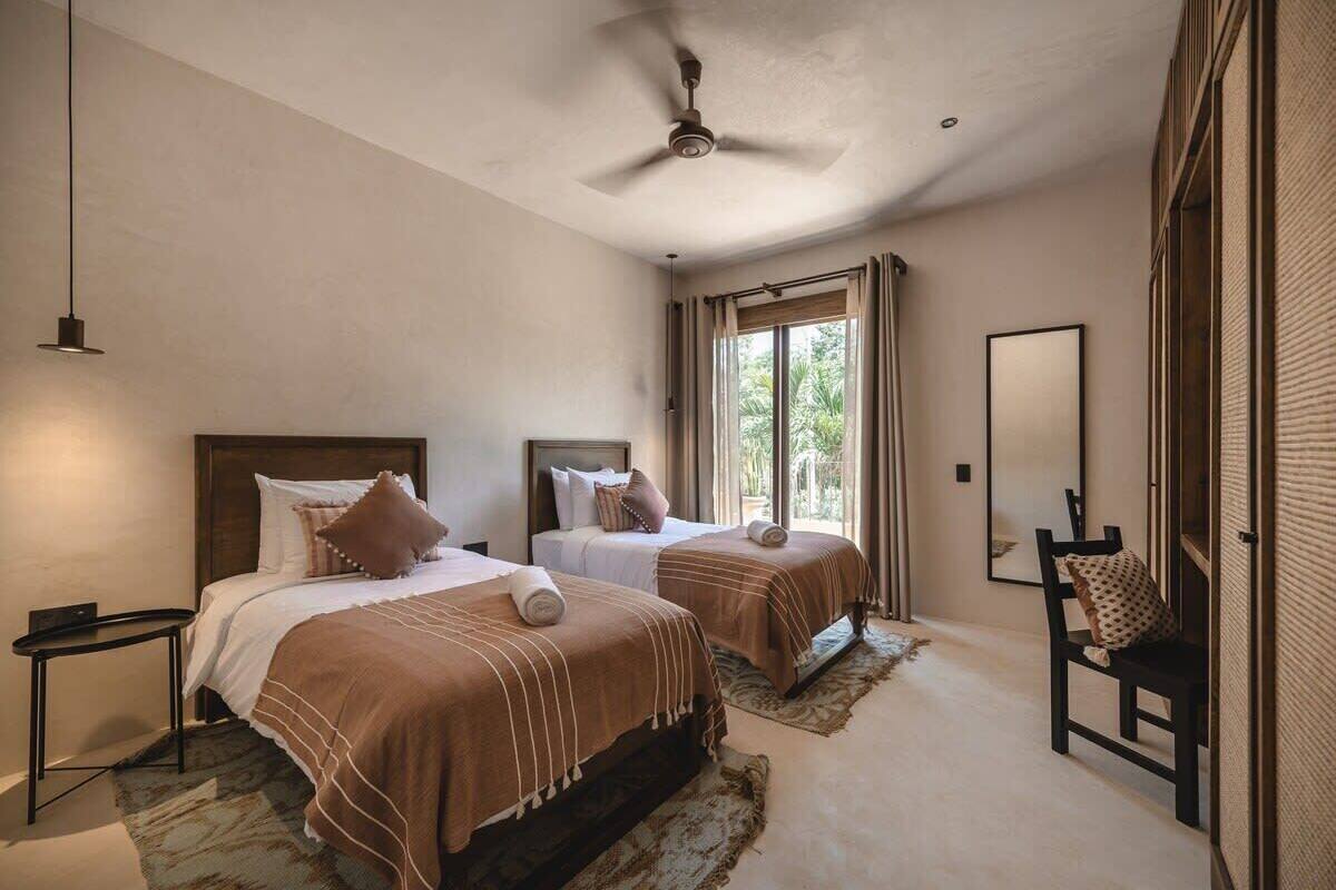 A bedroom with two beds, a ceiling fan, a window with curtains, a chair, and a mirror.