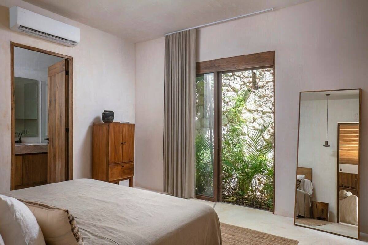 A bedroom with a bed, a wooden wardrobe, a large mirror, and a sliding glass door leading to a balcony with greenery.