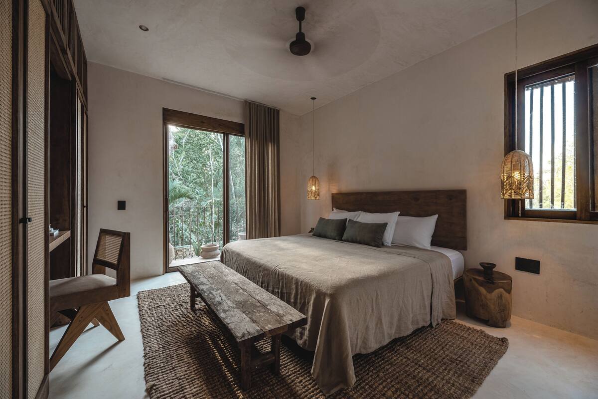 A bedroom with a large bed, wooden headboard, a bench, a window with curtains, and a hanging light fixture.