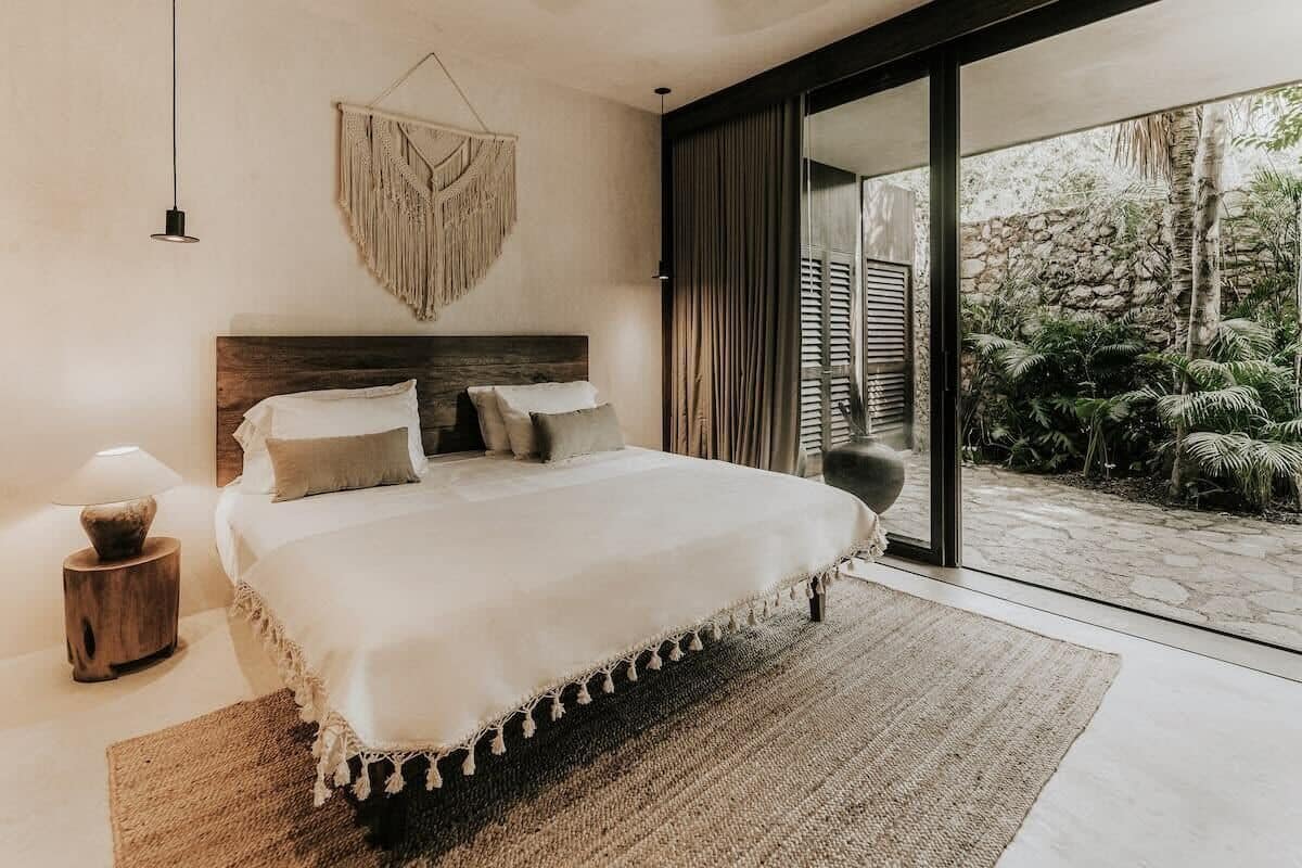 A bedroom with a large bed, a wooden headboard, a macrame wall hanging, and a view of a garden through a sliding glass door.