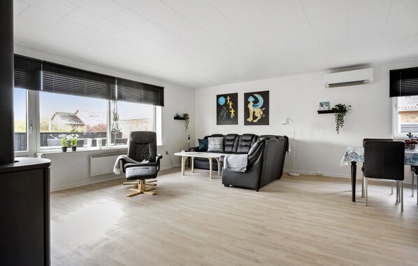 A spacious living room with a black leather sofa, a grey armchair, and a small table. There are two windows with blinds, a wall-mounted air conditioner, and a few wall art pieces.