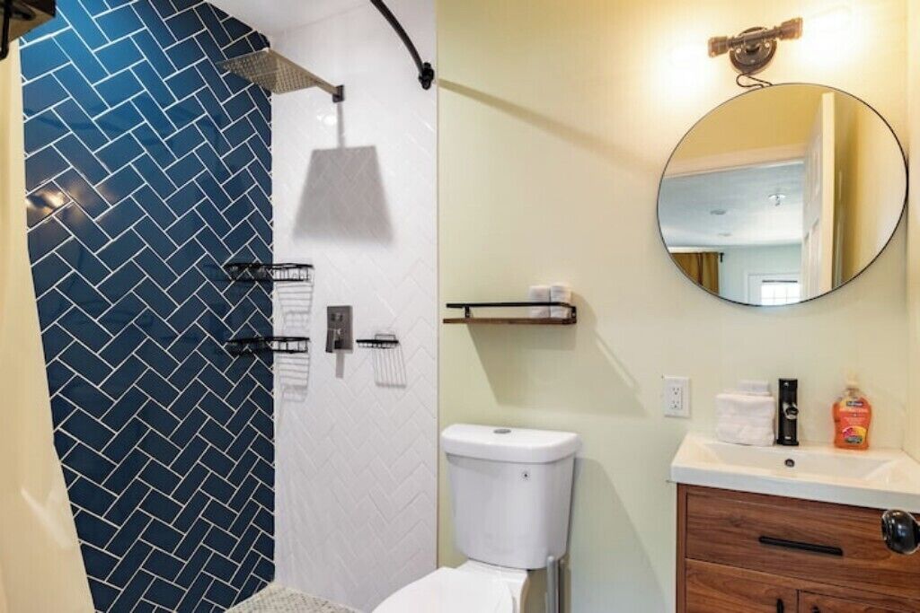 A bathroom with a blue herringbone tile shower, a white toilet, a wooden cabinet with a mirror, and a sink with toiletries.