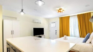 Habitación de hotel con una isla central grande, una cama con ropa blanca, un televisor y una ventana con cortinas amarillas.