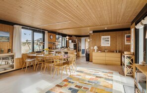 Ein geräumiger Raum mit Holzwänden und -decke, einem großen Esstisch mit Stühlen, einer Küchenzeile und Blick durch ein Fenster.
