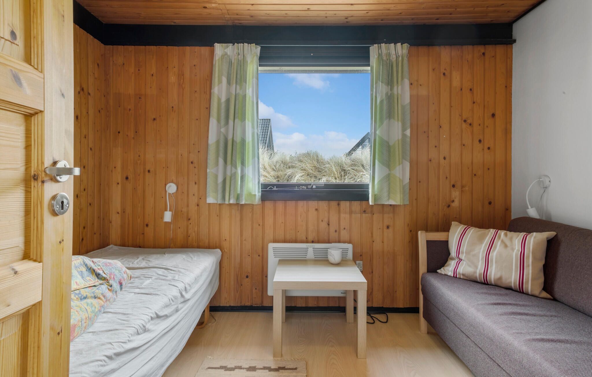 A small, cozy room with wooden walls, a window with curtains, a bed, a table, and a sofa.