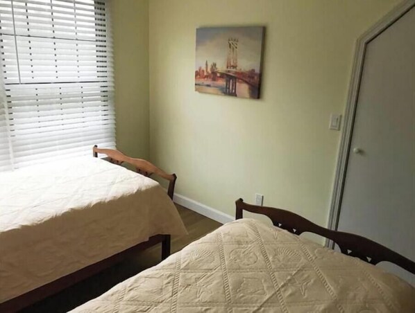 A bedroom with two beds, a window with blinds, a painting of a bridge, and a door.