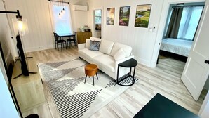 Ein modernes Wohnzimmer mit einem weißen Sofa, einem Couchtisch, einer schwarzen Stehlampe und einem schwarzen Beistelltisch. Es gibt eine Essecke mit Tisch und Stühlen, sowie eine Küchenzeile mit Schrank und Fenster.
