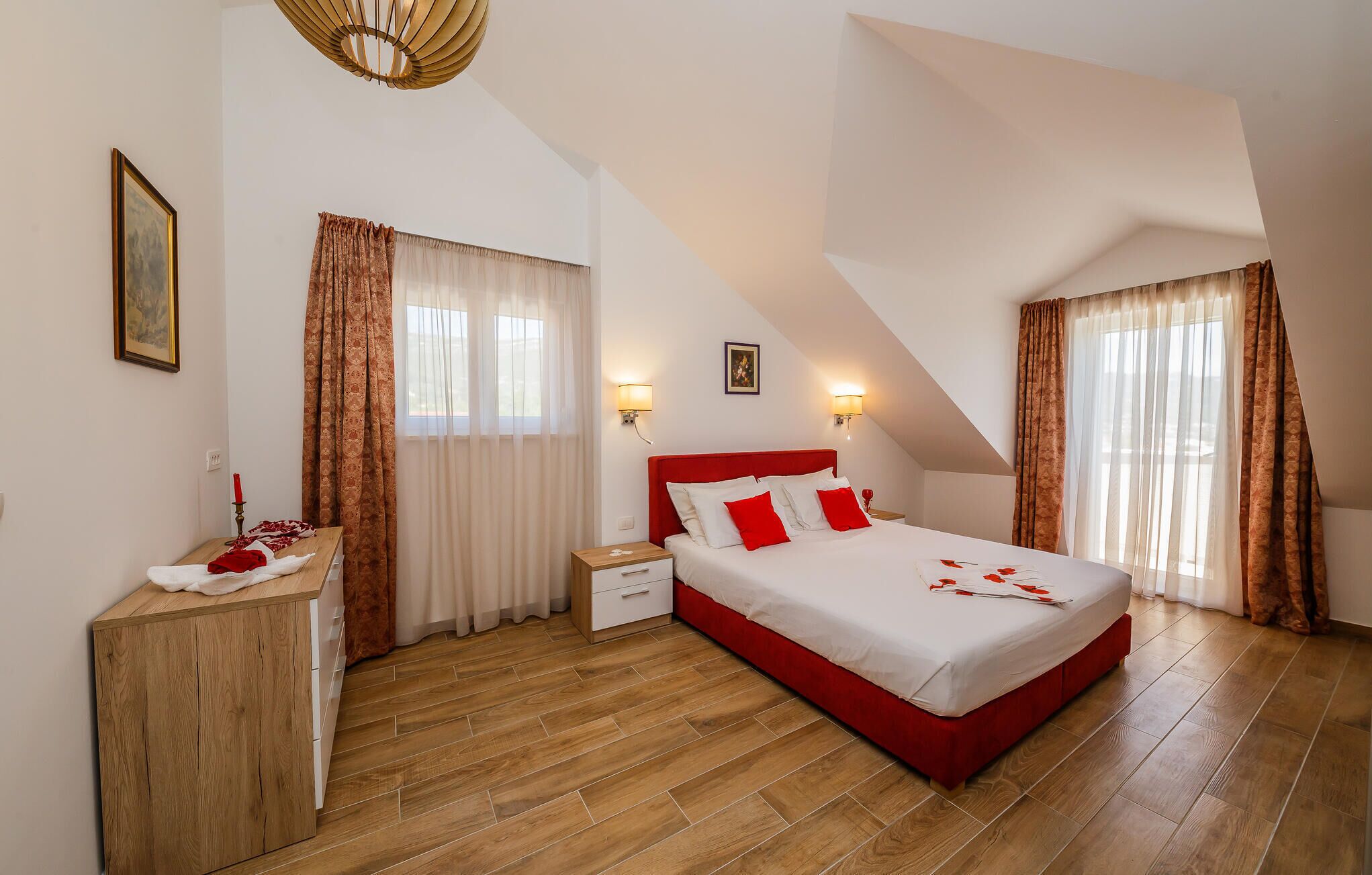 Un dormitorio con cama roja, suelo de madera, dos ventanas con cortinas, una mesita de noche y luces fijadas a la pared.