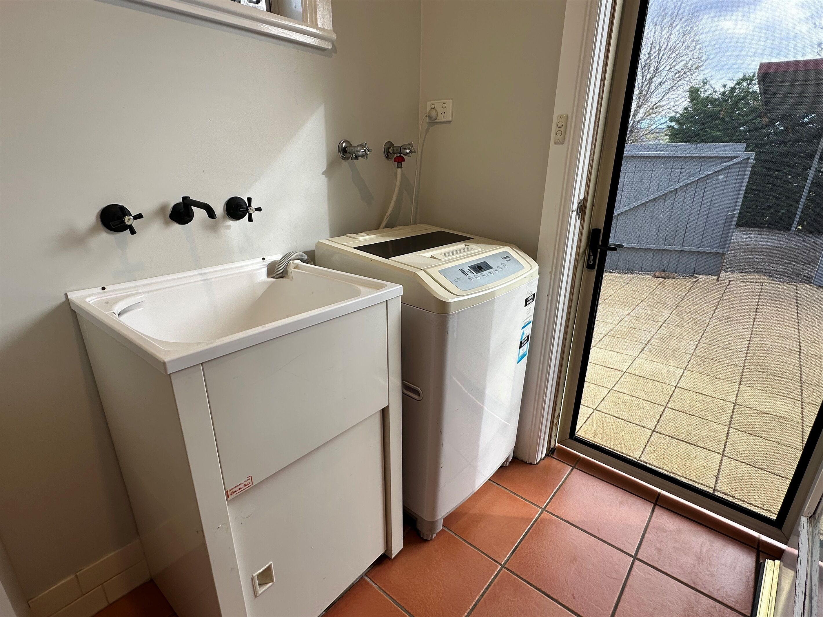 A laundry area with a sink and a washing machine, a tiled floor, and a door leading to an outdoor area with a fence.