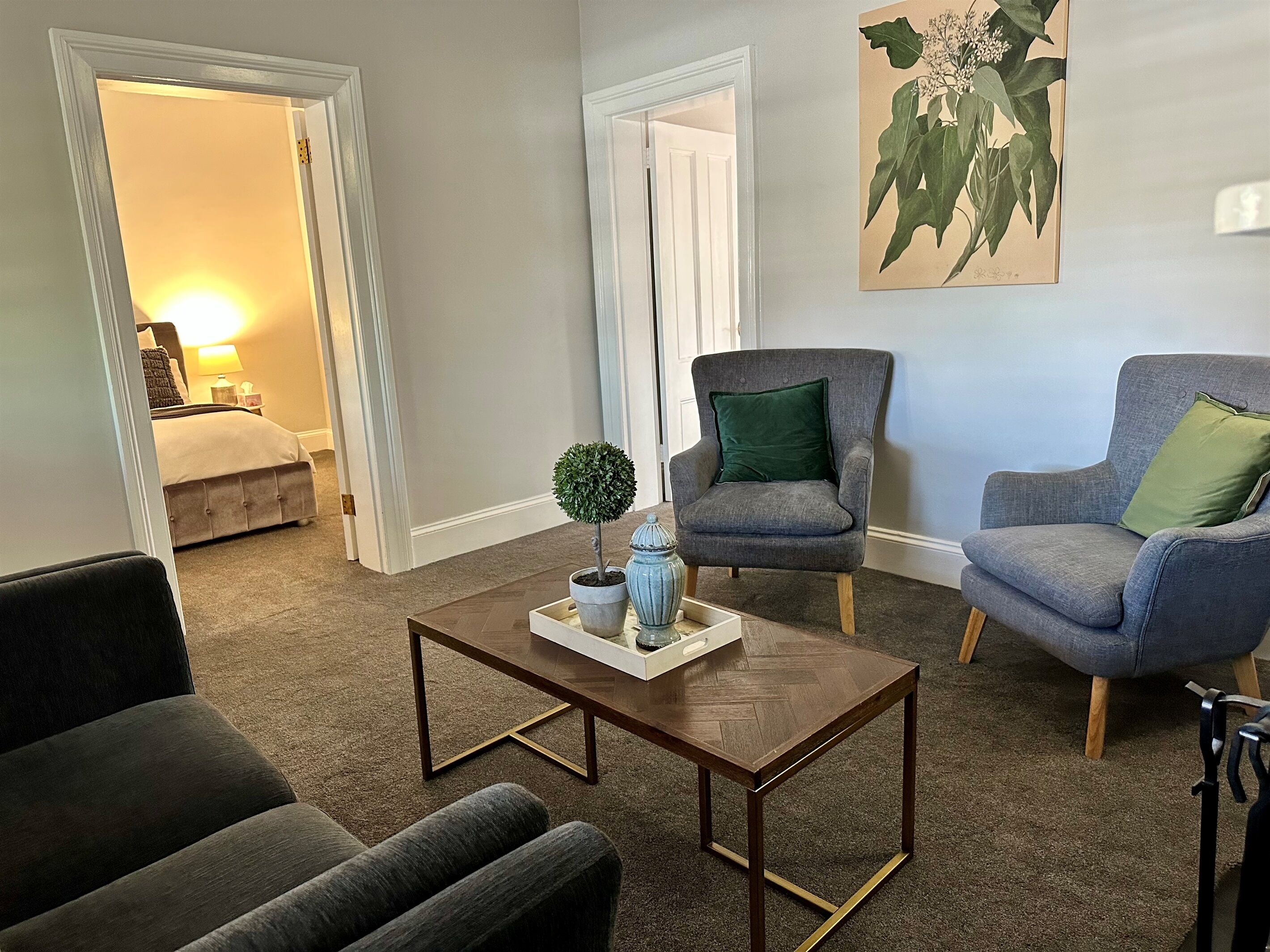 A living room with a dark brown herringbone coffee table, two grey armchairs with green cushions, and a small potted plant.