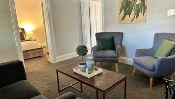 A living room with a dark brown herringbone coffee table, two grey armchairs with green cushions, and a small potted plant.