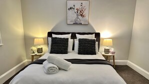 Ein ordentlich bezogenes Bett mit einem aufgerollten weißen Handtuch, zwei schwarzen, strukturierten Kissen und einem weißen Kopfteil. Es gibt zwei Nachttische, auf einem steht eine Lampe und ein kleiner Blumenstrauß.