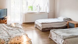 A bedroom with two single beds, a wooden floor, a radiator, a television on a wooden stand, and a window with white curtains.