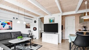 Un salon moderne avec un canapé, une table basse et un téléviseur. Il y a également un coin repas avec une table et des chaises. La pièce est dotée de poutres en bois au plafond et d’une grande fenêtre.