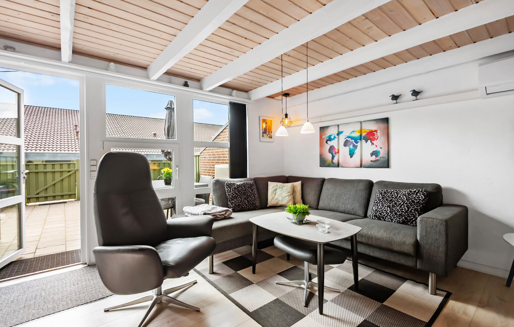 Un salon moderne avec un canapé gris, un fauteuil noir et une table basse. On y trouve deux lampes suspendues, une œuvre d’art représentant une carte du monde et une vue sur le jardin à travers de grandes fenêtres.