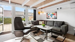 Un salon moderne avec un canapé gris, un fauteuil noir et une table basse. On y trouve deux lampes suspendues, une œuvre d’art représentant une carte du monde et une vue sur le jardin à travers de grandes fenêtres.