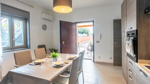 A modern kitchen with a dining area, featuring a wooden table set for four, a hanging light fixture, and a view through an open door to an outdoor area.