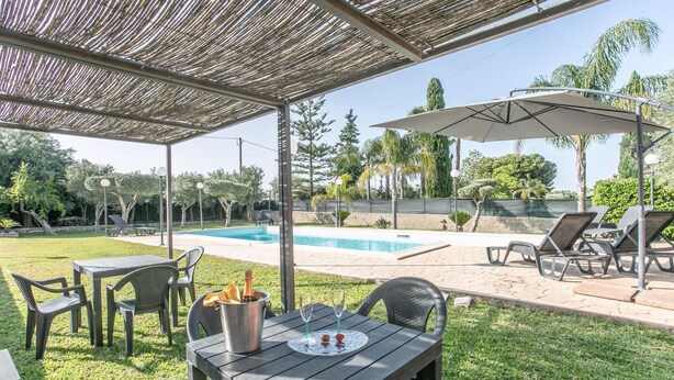 A covered outdoor area with a pool, lounge chairs, and a table with a bottle and glasses.