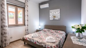 A bedroom with a bed, an air conditioner, a window with curtains, a bedside table with a lamp, and a wall with a flower painting.
