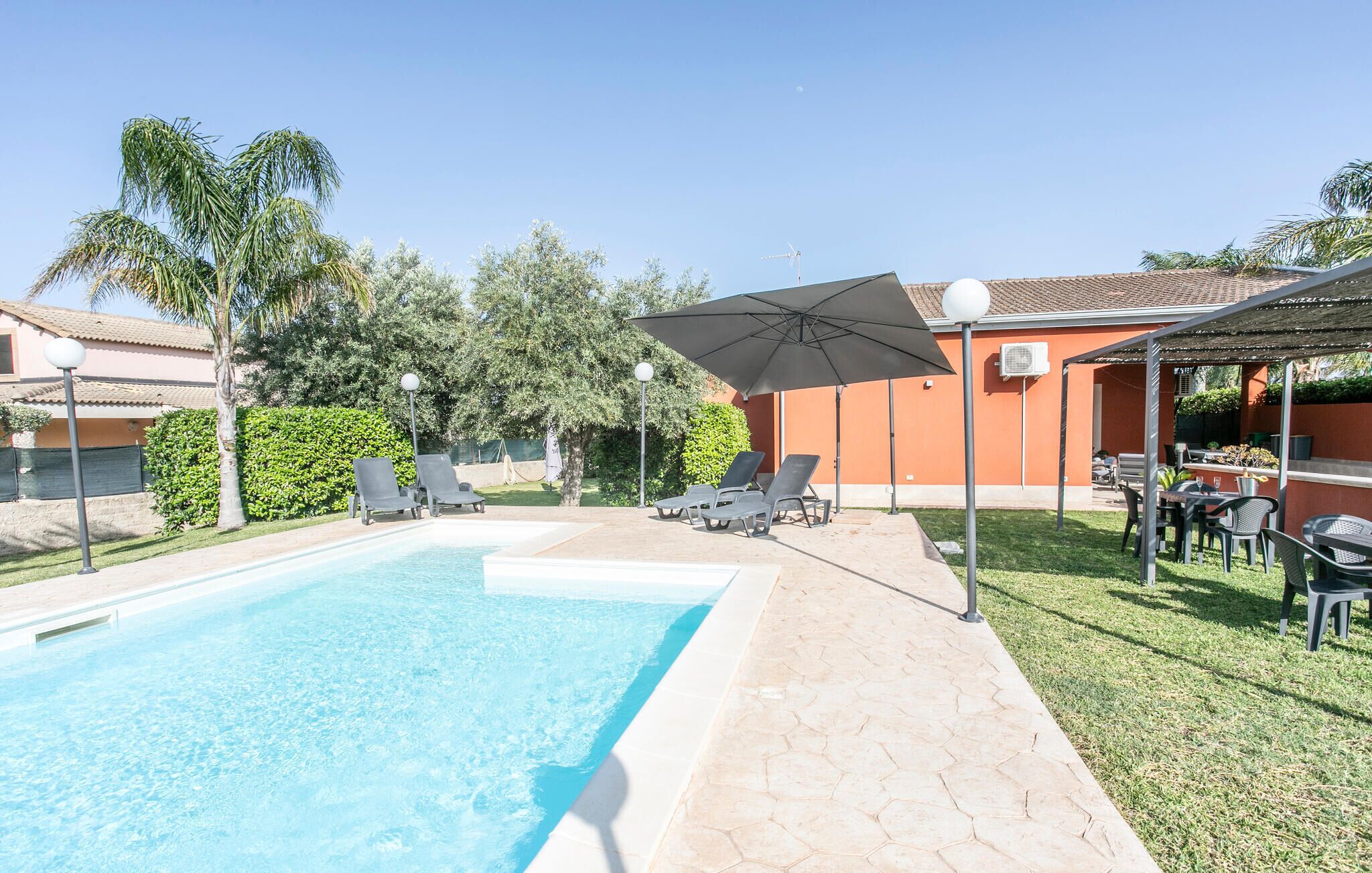 A swimming pool with lounge chairs and umbrellas, surrounded by greenery and a building with a tiled roof.