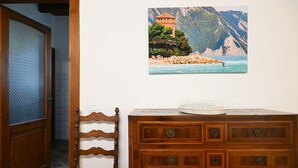 Uma cômoda de madeira com gavetas, uma cadeira com assento de palha e um quadro na parede retratando uma paisagem costeira.