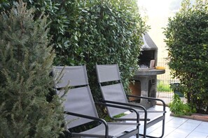 Two metal chairs with mesh backs and metal frames, positioned in front of a stone barbecue grill, surrounded by dense green foliage. - M424 - Marcelli, two-room apartment on the ground floor in a quiet area (Marcelli)