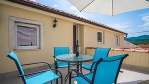 A balcony with a round table and four blue chairs, a window with a curtain, and a view of a tiled roof and hills. - Lovely apartment in Veli Brgud with WiFi (Veli Brgud)