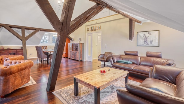 Un loft spacieux avec des poutres apparentes, un coin repas, des canapés en cuir et une cuisine à l’arrière-plan.