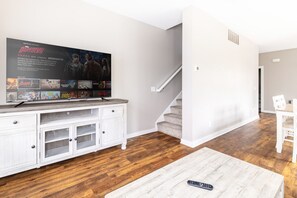 A living room with a TV mounted on a white cabinet, a staircase, and a dining area with a white table and chairs. - Luxury Downtown Townhome w/ Private Garage Unit 11 (Cleveland)