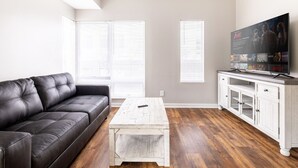 Una sala de estar con un sofá de cuero negro, una mesa de centro blanca y un televisor sobre un mueble blanco.