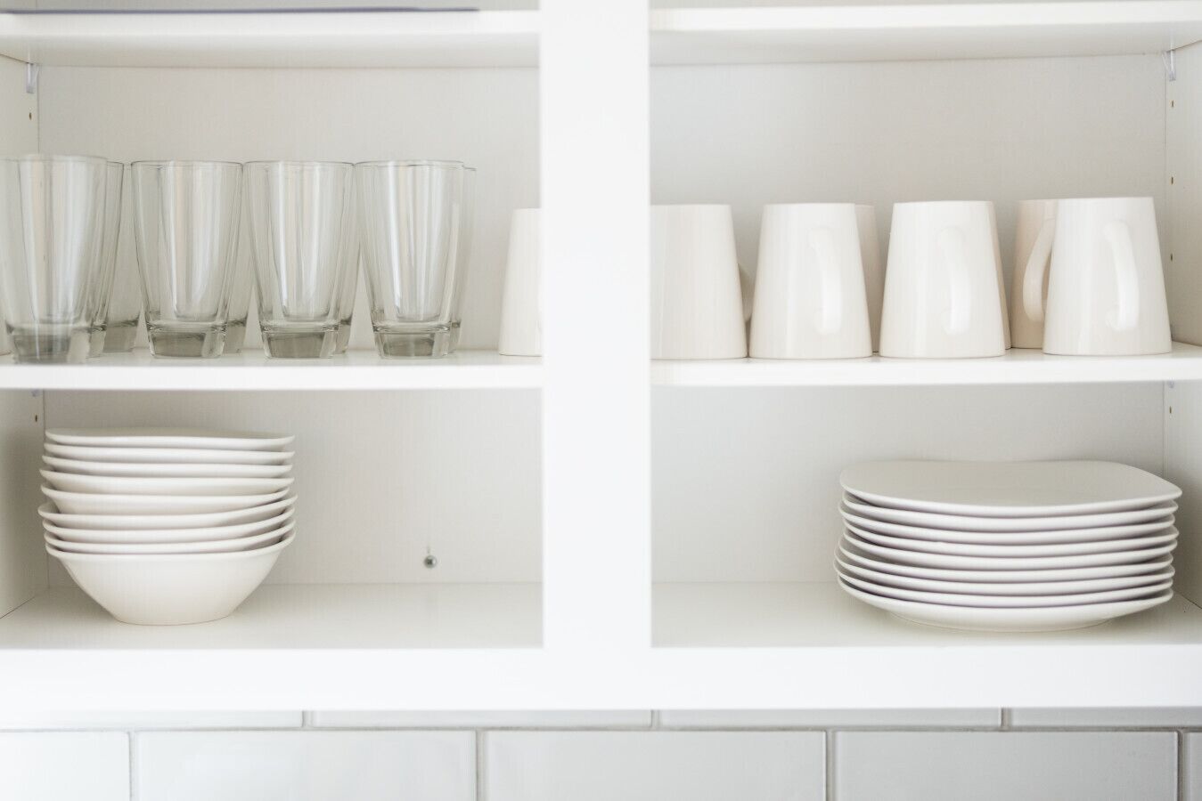 A kitchen with white cabinets and shelves. There are clear glasses, white cups, and stacked white plates.