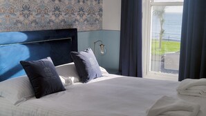 A hotel room with a bed, two pillows, a nightstand, a lamp, and a view of the ocean. - Kings' House (Douglas)
