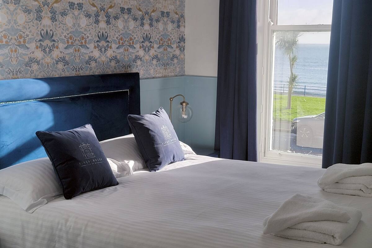 A hotel room with a bed, two pillows, a nightstand, a lamp, and a view of the ocean.