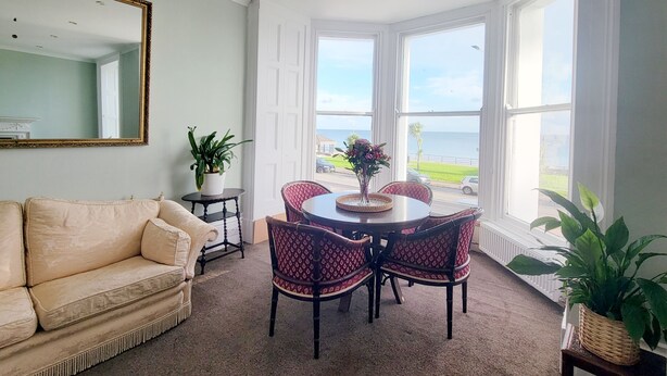 A room with a beige sofa, a dining table with four chairs, a large mirror, and a view of the ocean through large windows. - Kings' House (Douglas)