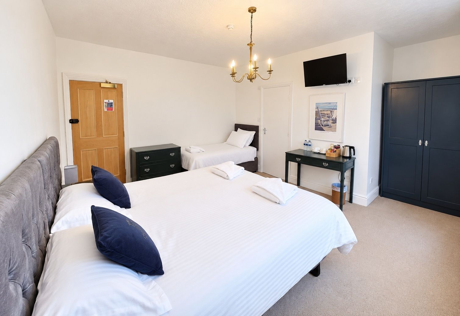 A hotel room with a large bed, a bedside table, a wardrobe, a television on the wall, and a chandelier.