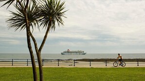 A coastal scene with a cruise ship in the distance, a cyclist on a path, and palm trees. - Kings' House (Douglas)