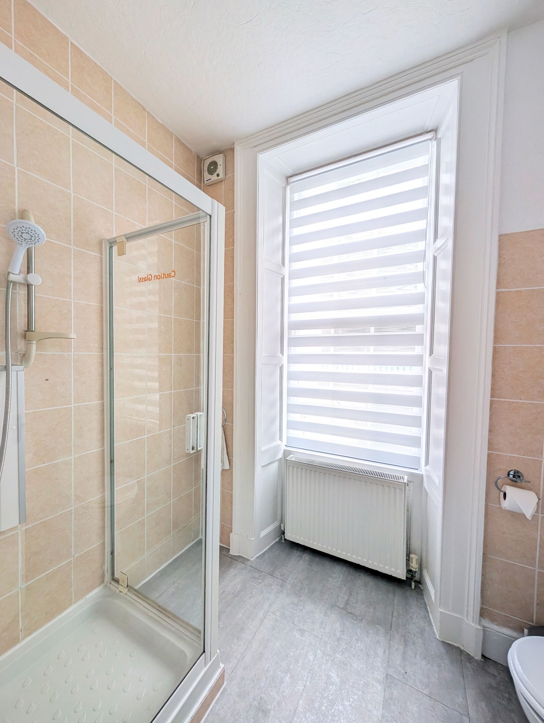 A bathroom with a glass shower enclosure, a toilet, and a window with blinds.