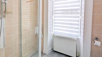 A bathroom with a glass shower enclosure, a toilet, and a window with blinds.